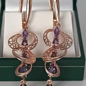 May include: A pair of rose gold earrings with a dangling design. Each earring features three oval filigree elements and several purple gemstones. The earrings are displayed in a green jewellery box.