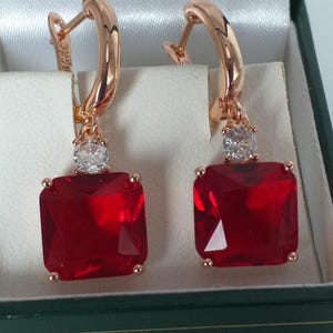 Könnte beinhalten: Ohrringe aus Roségold mit großen, quadratischen, rubinroten Edelsteinen. Jeder Ohrring hat einen kleineren, klaren, runden Stein über dem roten Edelstein. Die Ohrringe werden in einer weiß satinierten Schmuckschatulle präsentiert.