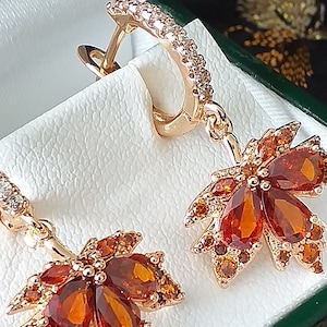 May include: A pair of gold earrings with a dangling leaf design. The leaves are set with orange and brown gemstones and are surrounded by smaller clear gemstones.