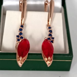 Könnte beinhalten: Ohrringe in Roségold-Optik mit einem tropfenförmigen, tiefroten Edelstein und einer Ansammlung kleiner blauer Edelsteine. Die Ohrringe werden in einer grünen Schmuckschatulle mit weißem Innenraum präsentiert.