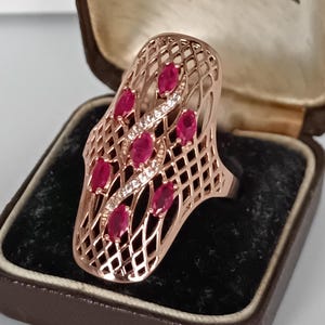 Op de afbeelding: Roségouden ring met een traliewerkontwerp, bezet met ovaal geslepen rode edelstenen en kleine heldere stenen. De ring wordt gepresenteerd in een vintage bruine sieradendoos met een zwart fluwelen interieur.