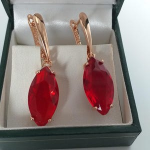 Op de afbeelding: Roségouden oorbellen met grote, druppelvormige, rode edelstenen. De oorbellen worden gepresenteerd in een donkergroene sieradendoos met een wit interieur. De edelstenen zijn gefacetteerd.