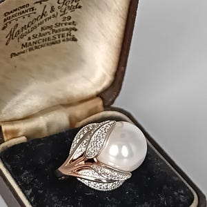 Puede incluir: Un anillo con una perla blanca grande, con un diseño de hojas, engastado en oro rosa y plata. El anillo se muestra en un joyero vintage marrón.