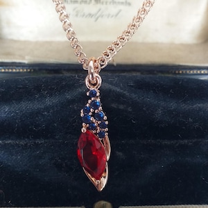 Könnte beinhalten: Halskette aus Roségold mit einem Anhänger mit einem großen roten Edelstein und kleineren blauen Edelsteinen. Der Anhänger hat eine Tropfenform. Die Halskette wird in einer Vintage-Schmuckschatulle präsentiert.