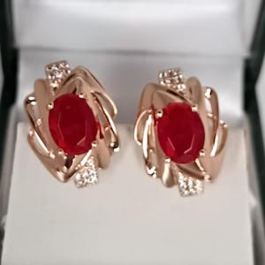 Könnte beinhalten: Ein Paar roségoldene Ohrringe, jeweils mit einem großen, ovalen roten Edelstein. Die Ohrringe haben ein dekoratives, wirbelndes Design mit kleinen, funkelnden Akzenten. Sie werden in einer weißen Schmuckschatulle präsentiert.