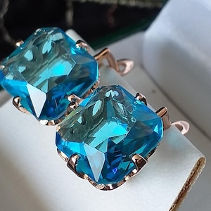 Può includere: Un paio di orecchini d'oro con accenti di pietre preziose blu. Le pietre preziose sono di forma quadrata e hanno un taglio sfaccettato.