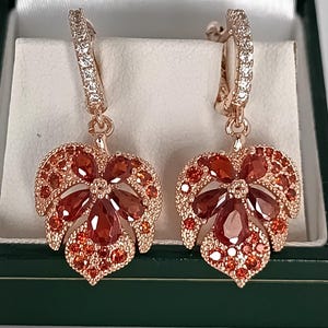 Peut inclure: Boucles d'oreilles en or rose avec des pendentifs en forme de feuilles ornés de pierres précieuses orange et rouges. Les créoles sont incrustées de petits cristaux transparents. Présentées dans un écrin vert.