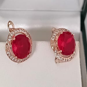 May include: Rose gold earrings, each with a large, oval, ruby-red gemstone. The earrings have a halo of small, clear crystals around the red stone. They are presented on a white surface.