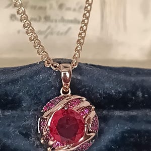May include: A rose gold-toned necklace featuring a round pendant with a large, faceted, deep red central stone. The pendant is surrounded by smaller, pavé-set red stones arranged in a swirling, textured pattern. The chain is a classic link style.