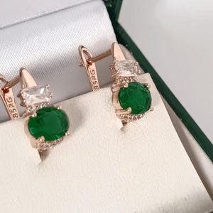 May include: Rose gold earrings with a lever back closure. Each earring features a large, oval, emerald-green stone, accented with small clear stones and a rectangular clear stone at the top. The earrings are displayed in a white jewelry box.