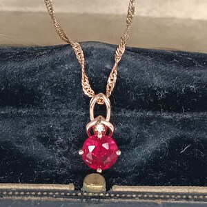 Könnte beinhalten: Halskette aus Roségold mit einer gedrehten Kette und einem Anhänger mit einem großen, runden, facettierten roten Edelstein. Ein kleiner Diamant akzentuiert die Oberseite des Anhängers. Die Halskette wird vor einem dunkelblauen Samthintergrund präsentiert.
