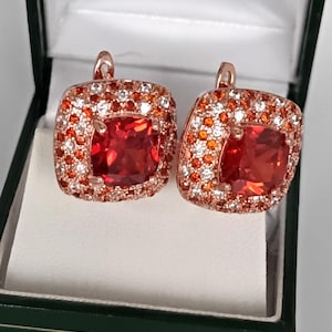 Pode incluir: Um par de brincos quadrados com uma grande pedra vermelha no centro, rodeada por pedras menores laranja e brancas. Os brincos são cravejados em metal de ouro rosa e apresentados numa caixa de joias verde.