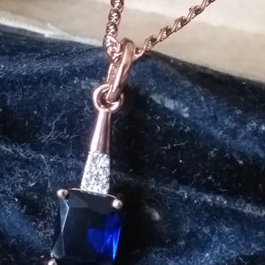 Puede incluir: Un collar de oro rosa con un colgante de piedra azul. El colgante está rodeado de piedras transparentes más pequeñas. El collar está en una caja de joyería.
