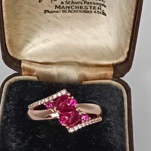 Op de afbeelding: Roségouden ring met een cluster van roze edelstenen en kleine witte stenen, gepresenteerd in een open zwarte fluwelen juwelendoos. De doos toont de tekst: "DIAMOND MERCHANTS. Hancock & Son (GOLDSMITHS) Ltd. 29 King Street & St. Ann's Passage MANCHESTER Phone: BLACKFRIARS 4512."