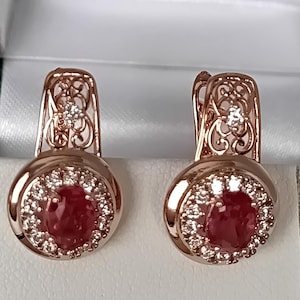 Könnte beinhalten: Ein Paar Ohrringe aus Roségold mit einem oval geschliffenen roten Edelstein in der Mitte, umgeben von einem Kranz aus kleinen klaren Steinen. Die Ohrhaken zeigen ein filigranes Metallmuster mit einem kleinen klaren Stein an der Spitze.
