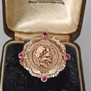 Puede incluir: Un anillo de oro rosa con un motivo de moneda central con un retrato de perfil, rodeado por un borde festoneado engastado con pequeñas piedras blancas y cuatro rubíes rojos redondos en los puntos cardinales.
