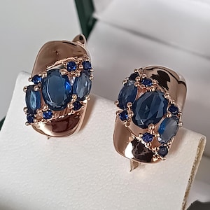 Può includere: Orecchini in oro rosa con un gruppo di pietre preziose blu ovali e rotonde. Gli orecchini sono esposti in un portagioie bianco, evidenziando il loro design e colore.