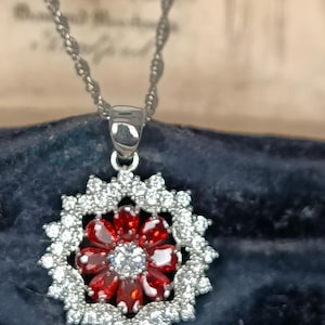 Könnte beinhalten: Eine silberfarbene Anhängerkette mit einem zentralen Blumendesign aus roten, ovalen Steinen, die einen klaren, runden Mittelstein umgeben. Die Blume ist von einem gewellten Rand aus kleinen, klaren, runden Steinen auf einer silberfarbenen Kette umrahmt.