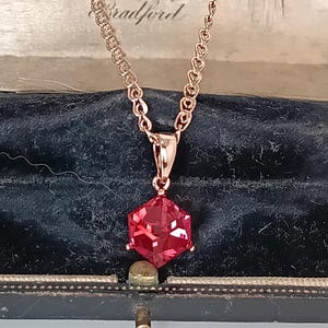 May include: A rose gold necklace featuring a red, faceted gemstone pendant. The pendant is a geometric shape, suspended from a delicate chain. The necklace is displayed in a vintage jewellery box.