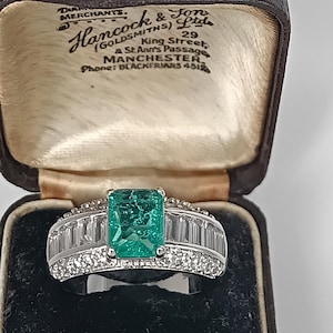 Peut inclure: Bague en argent ornée d'une grande pierre verte taillée en émeraude au centre, entourée de petits diamants clairs. La bague est présentée dans un écrin vintage avec le texte "Hancock & Son Ltd, Goldsmiths, Manchester".