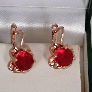 Puede incluir: Un par de pendientes de oro rosa con un diseño en forma de corazón. Cada pendiente presenta una piedra preciosa grande, redonda y de color rojo brillante. Los pendientes se exhiben en una caja de joyería blanca.