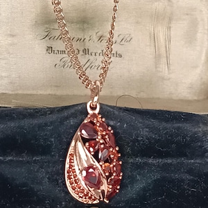 Könnte beinhalten: Halskette mit roségoldfarbenem Anhänger, der eine tropfenförmige Form hat. Der Anhänger ist mit roten Edelsteinen und einem detaillierten Design verziert. Die Halskette wird in einer Vintage-Schmuckschatulle präsentiert.