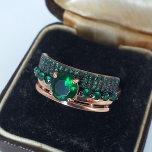 Op de afbeelding: Een roségouden ringenset met een grote, ronde, smaragdgroene edelsteen en kleinere groene stenen. De set wordt gepresenteerd in een zwarte fluwelen doos, die de glans en het ontwerp van de sieraden benadrukt. Het is een opvallend sieraad.