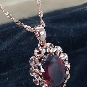 May include: A rose gold necklace with a large, oval, deep red gemstone pendant. The pendant is surrounded by a decorative, scalloped frame. The chain is a delicate rope style. The jewelry is displayed against a dark background.