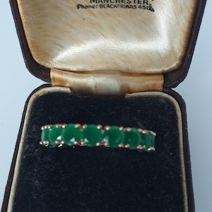 Puede incluir: Un anillo de oro con piedras preciosas verdes de corte ovalado, exhibido en un joyero vintage. La caja es marrón con un forro de satén color crema. El texto "Hancock & Son" es visible.