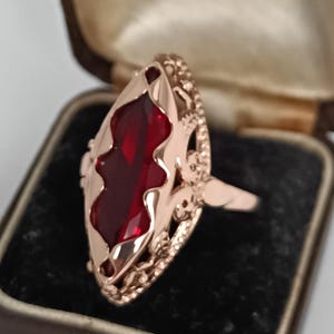 May include: A rose gold ring with a large, marquise-cut, deep red gemstone. The ring features intricate filigree detailing around the gemstone and along the band. The ring is displayed in a vintage brown jewelry box with a black velvet interior.