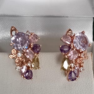 May include: Rose gold earrings with clusters of gemstones in shades of lavender, pink, and purple. The earrings have a delicate design with butterfly accents and small white stones. The gemstones are various shapes and sizes.