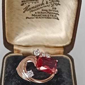 May include: A rose gold ring featuring a large rectangular red gemstone and three small clear round stones. The ring is displayed inside a brown vintage jewelry box with cream-colored lining and text reading Diamond Merchants Hancock & Son.
