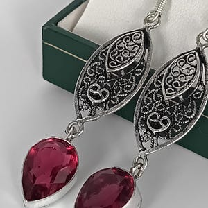 May include: Silver filigree earrings with teardrop-shaped, ruby-red gemstones. The earrings have an oval filigree design with detailed patterns and a hook closure. The gemstones are set in a silver bezel.