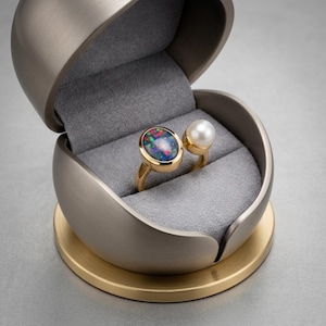 Könnte beinhalten: Ein goldener offener Ring mit einem großen ovalen schwarzen Opal mit leuchtend roten und grünen Blitzen an einem Ende und einer einzelnen runden weißen Perle am anderen, präsentiert in einer kugelförmigen Schmuckschatulle aus gebürstetem Metall.