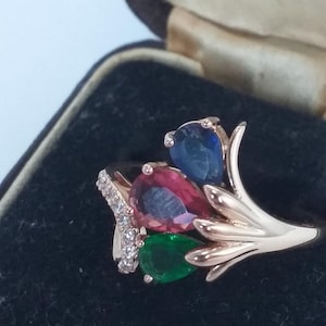 Könnte beinhalten: Ein goldfarbener Ring mit drei tropfenförmigen Edelsteinen in Blau, Rot und Grün. Der Ring hat ein florales Design mit einem Band aus kleinen, klaren Steinen. Der Ring ist vor einem dunklen Hintergrund platziert.
