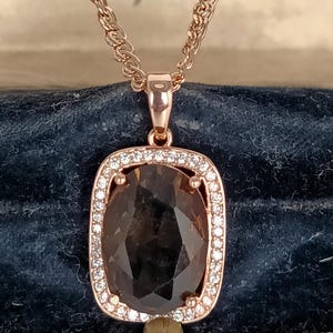 Puede incluir: Collar con colgante de oro rosa con una gema ovalada grande, de color marrón oscuro. La gema está rodeada por un halo de pequeños cristales transparentes. La cadena es de color oro rosa.