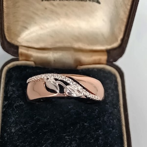 May include: A rose gold ring with a wave design, featuring a line of small, clear sparkling stones. The ring is presented in a vintage brown jewellery box with a black velvet interior.