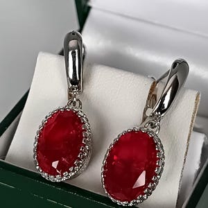 Pode incluir: Um par de brincos pendentes em tom prateado com grandes pedras vermelhas ovais facetadas. Cada pedra está engastada numa moldura de metal prateado decorativa e recortada. Os brincos têm um fecho de alavanca prateado polido.