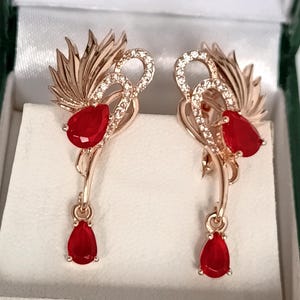 May include: Rose gold earrings with a leaf design and sparkling details. Each earring has a large, teardrop-shaped, red gemstone and a smaller, dangling red gemstone. The earrings are presented in a white box with a green border.