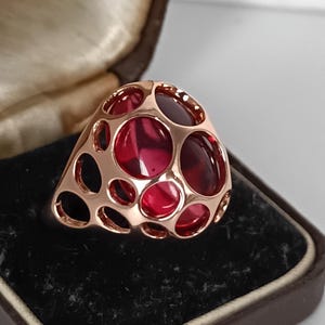 May include: Rose gold ring with a unique openwork design, featuring large, round, red gemstones. The ring is displayed in a vintage brown jewelry box with a black velvet interior. The ring's design includes multiple circular openings.