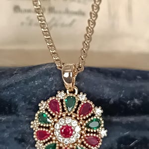 Op de afbeelding: Goudkleurige hangerketting. De cirkelvormige hanger heeft een centrale rode edelsteen, omringd door witte stenen. Afwisselend rode en groene druppelvormige edelstenen sieren de hanger. De ketting heeft een fijne schakel.
