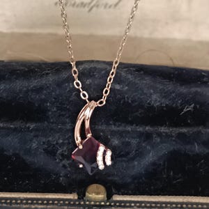 Könnte beinhalten: Eine roségoldene Halskette mit einem Anhänger mit einem dunkelroten quadratischen Edelstein und einem geschwungenen Akzent. Der Anhänger ist mit kleinen, funkelnden weißen Steinen verziert. Die Halskette wird in einer Vintage-Schmuckschatulle präsentiert.