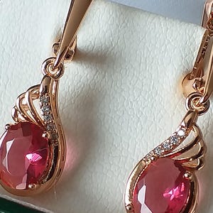 May include: A pair of rose gold earrings with a teardrop-shaped red gemstone and a delicate design. The earrings have a small, clear stone accent on the side.