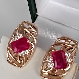 Op de afbeelding: Een paar roségouden oorbellen met een rechthoekige, dieprode edelsteen in het midden. De oorbellen hebben een open filigraan ontwerp met kleine, witte accentstenen. De oorbellen worden gepresenteerd in een witte sieradendoos.