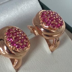 Op de afbeelding: Een paar roségouden oorbellen met een cluster van kleine, helder roze edelstenen in een ovale vorm. De oorbellen worden gepresenteerd in een witte sieradendoos, die hun gepolijste afwerking en gedetailleerde ontwerp benadrukt.