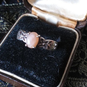 Könnte beinhalten: Ein silberner Ring mit einem rosafarbenen ovalen Edelstein und floralen Designs. Der Ring befindet sich in einer schwarzen Samt-Ringbox.