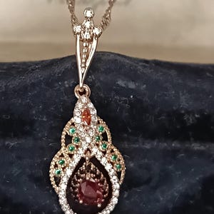 Può includere: Una collana con pendente in oro rosa con un design a forma di goccia. Il pendente presenta una grande gemma rossa rotonda al centro, circondata da gemme bianche e verdi più piccole. La collana ha una catena delicata.