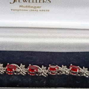Pode incluir: Uma pulseira de tom prateado com uma série de pedras vermelhas redondas e facetadas alternadas com pequenas pedras transparentes. A pulseira é apresentada dentro de uma caixa de joias forrada de veludo azul escuro com o texto Harbour Jewellers.