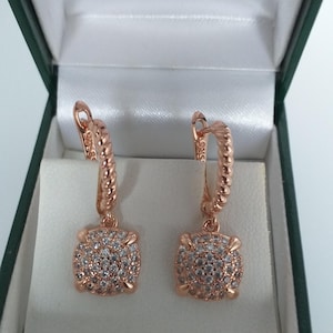 Peut inclure: Boucles d'oreilles de couleur or rose avec un design carré incrusté de cristaux. Les boucles d'oreilles sont présentées dans un écrin vert foncé ouvert avec un intérieur blanc. Les boucles ont une boucle texturée et un pendentif carré recouvert de cristaux.