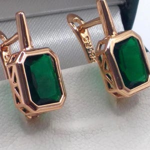 May include: A pair of gold earrings with emerald green gemstones. The earrings have a square shape and are set in a delicate gold frame.